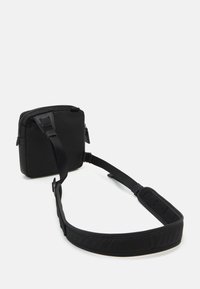 Kompaktowa czarna torba na ramię z regulowanym, wyściełanym paskiem i zapięciem na suwak, leżąca na białej powierzchni.