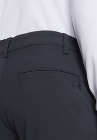 Pantalon gris foncé avec une texture lisse, présentant une poche arrière, des détails cousus et un logo Under Armour noir près de la taille.