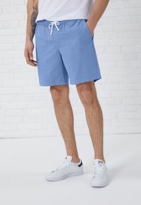 Pantaloni da bagno azzurri chiari con vita elasticizzata e cordoncino, dotati di tasche laterali e una texture di tessuto liscia. Indossati con sneaker bianche.