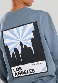 Sudadera azul con un gran gráfico negro de un horizonte urbano, rayos de fondo azul claro y el texto "LOS ÁNGELES" debajo.