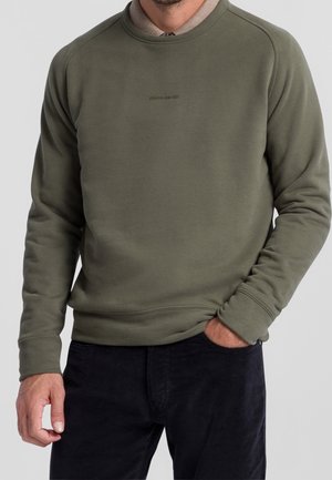 Hombre con suéter de cuello redondo color verde oliva sobre camisa beige con cuello y pantalones negros, con una mano en el bolsillo.