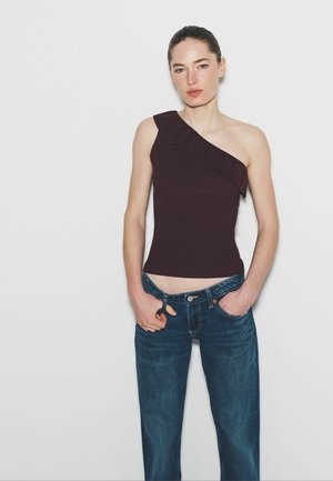 Woman wearing a sleeveless, one-shoulder dark purple top with ruffle detail and blue jeans, standing with hands in pockets against plain background.