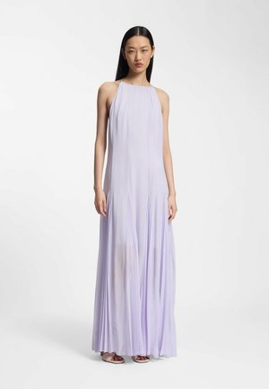 Kvinde med langt sort hår iført en ærmeløs, gulvlængde, lys lavendelfarvet plisseret kjole og åbne hæle, stående foran en ensfarvet baggrund.
