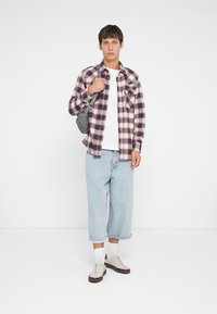 Camisa de franela a cuadros en púrpura y blanco, camiseta blanca, pantalones de mezclilla anchos en azul claro, mochila gris y zapatillas beige con suelas oscuras.