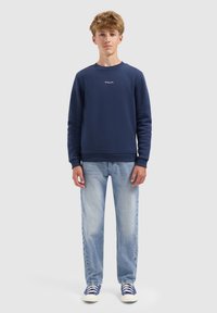 Tienerjongen staat met zijn gezicht naar voren gekeerd, draagt een marineblauwe sweater, lichtblauwe jeans en blauwe sneakers tegen een effen grijze achtergrond.