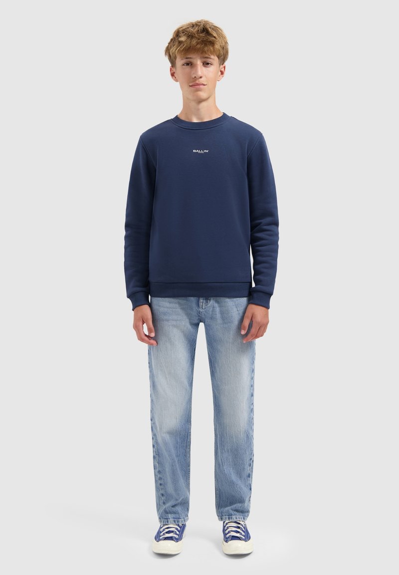 Tienerjongen staat met zijn gezicht naar voren gekeerd, draagt een marineblauwe sweater, lichtblauwe jeans en blauwe sneakers tegen een effen grijze achtergrond.