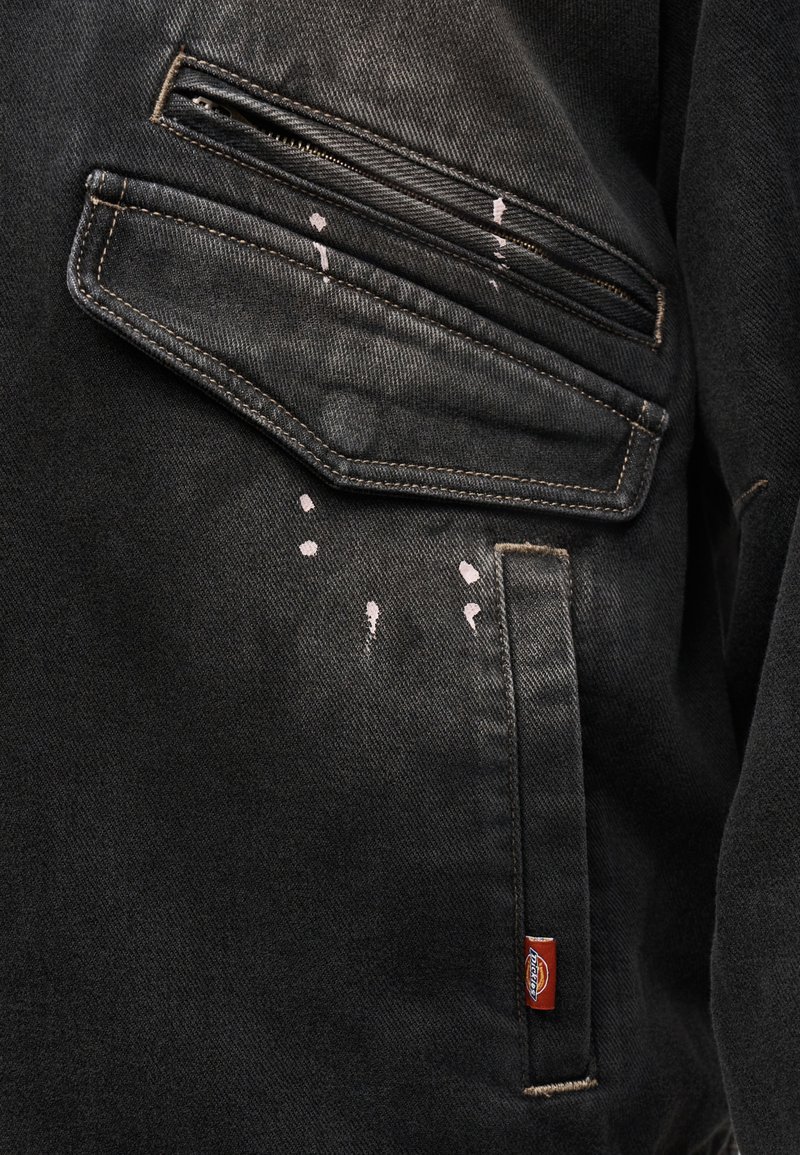 Sezione di giacca in denim nera con tasche con cerniera e a patta, con piccole macchie di vernice bianca e un'etichetta del marchio in tessuto rosso.