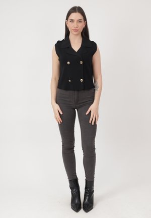 Młoda kobieta stoi, ubrana w bezrękawnik czarny top z podwójnym rzędem guzików, ciemnoszare obcisłe dżinsy, czarne botki na obcasie oraz z widocznym tatuażem na ramieniu.