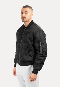 Giacca bomber nera con una finitura satinata liscia, chiusura frontale con zip e polsini a costine. Presenta tasche laterali e una tasca con zip sulla manica.