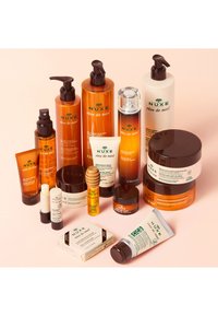 Kolekcja kosmetyków do pielęgnacji skóry NUXE Rêve de Miel z butelkami w odcieniach bursztynu i bieli, w różnych kształtach, etykietami z zielonym tekstem oraz drewnianymi aplikatorami.