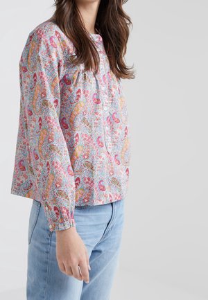 Frau trägt eine langärmelige Bluse mit buntem Paisley-Muster und hellblaue Jeans, steht vor einem einfarbigen hellgrauen Hintergrund.