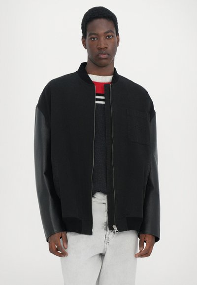 Black bomber jacket with leather sleeves, zip closure, side pocket, and ribbed hem. Worn over a striped knit sweater.