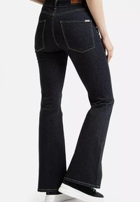 Kobieta ubrana w ciemnoniebieskie dżinsy w kształcie dzwonów i czarne sneakersy z białymi podeszwami, pokazana od tyłu na białym tle.