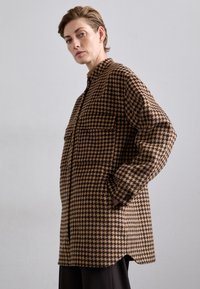 Bruin en zwart houndstooth-patroon hemdjack met een knoopsluiting, twee borstzakken en lange mouwen. Gestructureerde stof.