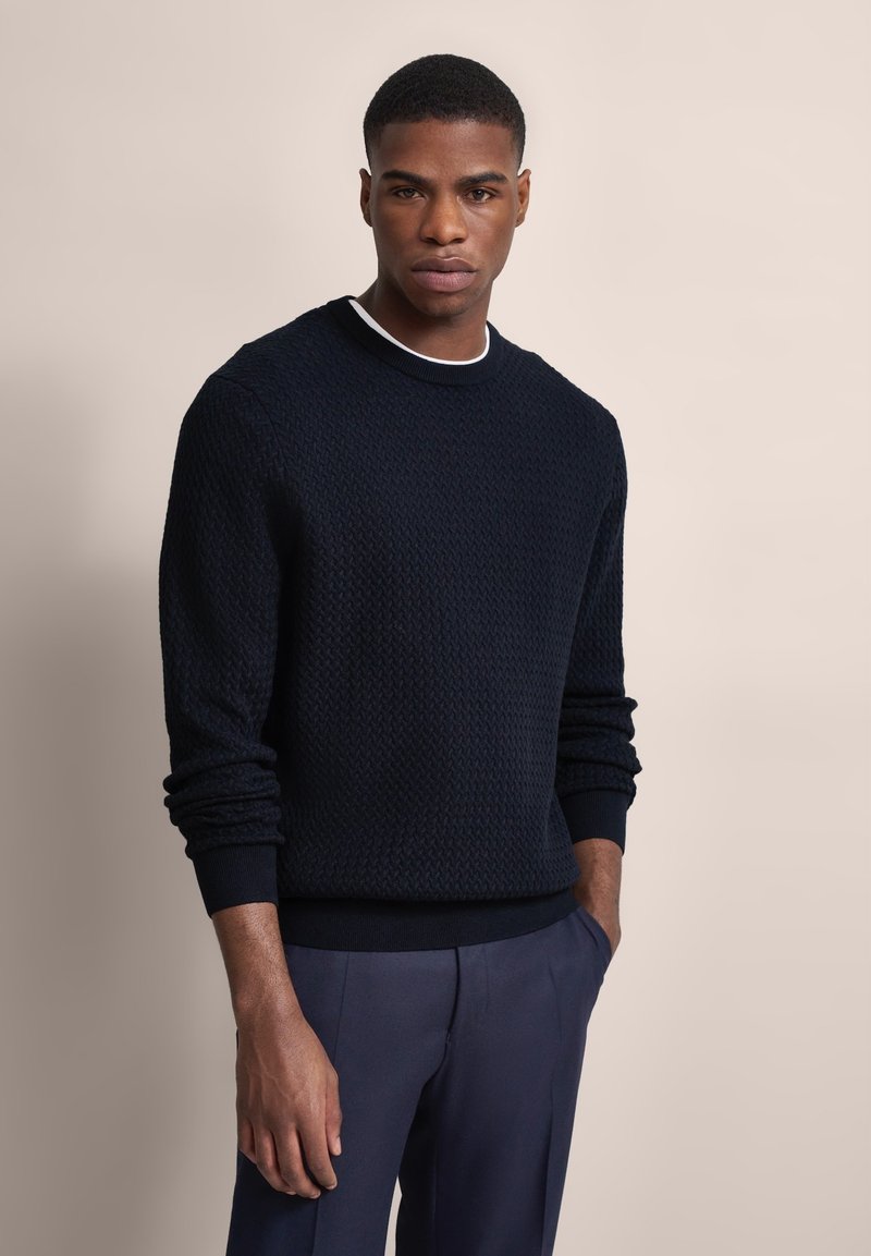 Marinepullover mit strukturiertem Strickmuster, langen Ärmeln und gerippten Bündchen sowie Saum, getragen über einem weißen Kragenhemd und kombiniert mit grauen Hosen.