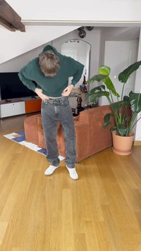 Zelená mikina s kapucňou, šedé džínsy a biele tenisky; stojí na drevenej podlahe pri kvetináči a textúrovanej oranžovej pohovke. Neutrálne pozadie.