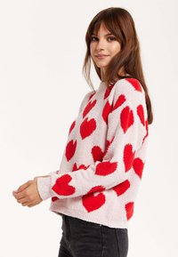 Crewneck sweater in light pink with raised red heart patterns. Soft texture, long sleeves, slightly cropped fit, and ribbed cuffs.