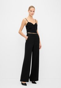 Top noir court avec de fines bretelles et un décolleté en cœur, associé à un pantalon noir à jambes larges avec des broderies en dentelle sur les côtés.