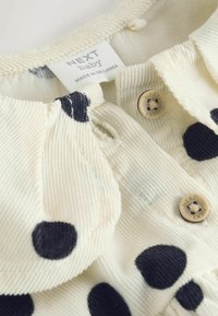 Gaiši krēmkrāsas kordurova audums ar lieliem melniem polka punktiem, ar zelta pogu aizdari un etiķeti "NEXT baby."