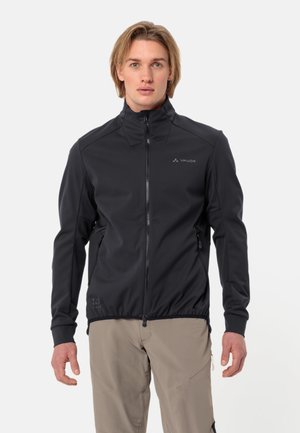 Giovane uomo con capelli biondi che indossa una giacca nera Vaude con cerniera e pantaloni beige, in piedi davanti a uno sfondo bianco semplice.