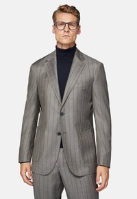 Blazer rayé gris avec revers crantés et deux boutons. Il possède une poche poitrine et des poches latérales. Porté par-dessus un col roulé noir.