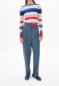 Person som bär en långärmad tröja med flerfärgade ränder, blå högmidjade byxor och blå slip-on skor mot en vit bakgrund.