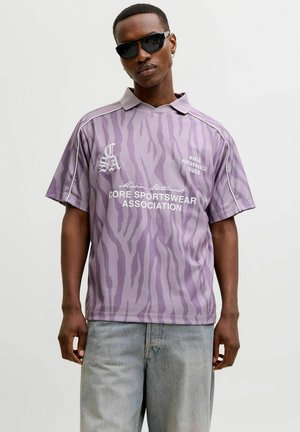 Homme portant des lunettes de soleil noires et un polo ample lilas à imprimé zèbre avec un texte blanc, ainsi qu'un jean bleu clair, posant devant un fond uni.