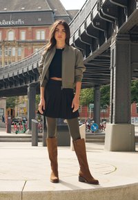 Olive bomber jacket over black crop top, black pleated skirt, olive leggings, and brown knee-high boots with block heels, standing outdoors.