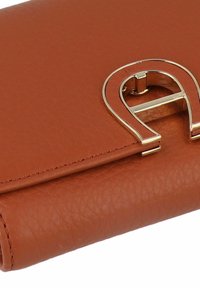 Brown leather wallet with a textured finish, featuring a shiny gold clasp shaped like an "A." Minimal stitching detail along the edges.