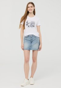 T-shirt bianca in cotone con grafica di un cane e testo, abbinata a una gonna di jeans azzurri leggeri e sneakers bianche, con orlo dritto.