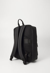 Zaino nero con schienale in rete testurizzata, spallacci imbottiti e finitura liscia, completo di maniglia superiore per un facile trasporto. Compartimento con zip visibile.