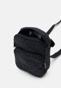 Petite sac à bandoulière noir avec poche zippée à l'avant et bandoulière réglable, affichant un motif subtil de logo trefoil Adidas et une étiquette à l'intérieur.