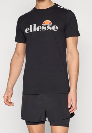 Homme portant un T-shirt noir ellesse et un short de sport superposé noir, se tenant contre un fond clair uni.
