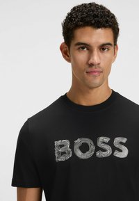 Musta puuvillapaita, jossa on teksturoitu valkoinen "BOSS" -grafiikka edessä. Klassinen pyöreä kaula-aukko ja lyhyet hihat. Siisti muotoilu.
