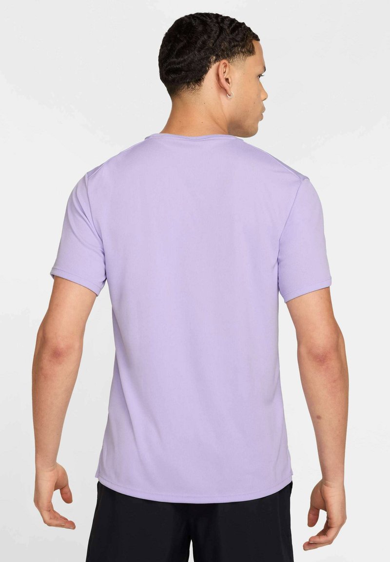 Homme aux cheveux courts et bouclés, portant une chemise lavande à manches courtes et un pantalon noir, de dos, debout devant un fond blanc uni.