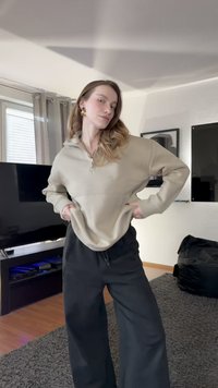 Sudadera con cremallera beige de corte holgado y bolsillo tipo canguro, combinada con pantalones de chándal negros sueltos. Habitación con suelo de madera y alfombra oscura.