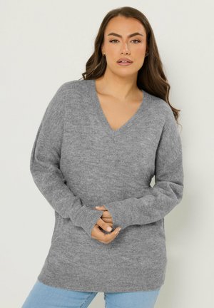 Kvinde iført en løs grå V-hals sweater og lyseblå jeans, stående mod en ensartet lys baggrund med hænderne foldet.