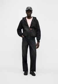 Μαύρο τζιν μπουφάν με φερμουάρ και κουκούλα, συνδυασμένο με μαύρο τζιν παντελόνι. Το μοντέλο φοράει ροζ πουκάμισο και μαύρα αθλητικά παπούτσια, ολοκληρωμένα με σκούφο σε σκούρο χρώμα.