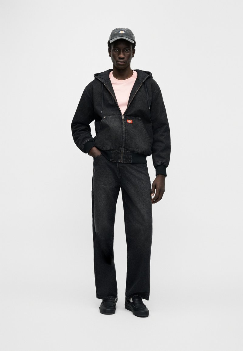 Zwarte spijkerjasje met ritssluiting en capuchon, gecombineerd met zwarte jeans. Model draagt een roze shirt en zwarte sneakers, afgewerkt met een donkere pet.