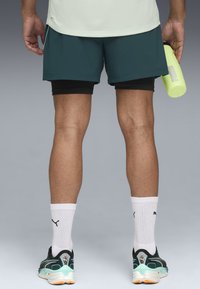 Persoon van achter met een teal korte broek over zwarte leggings, witte Puma sokken, teal sneakers, en een gele Puma waterfles in de rechterhand.