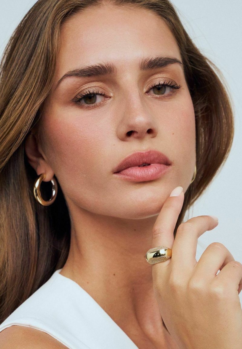 Guldring med en polerad yta och texturerad band, bärs på ett finger. Modellen har medelbrunt hår och en neutral sminkpalett. Bakgrunden är ljus.