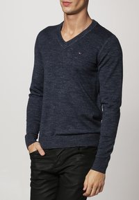 Homme portant un pull à col en V bleu marine ajusté et un pantalon noir, une main dans la poche, debout devant un fond clair uni.