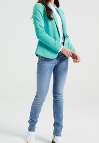 Turkos blazer med en figurnära design, kombinerad med ljusblå slim fit jeans. Vita knappar och manschetter ger kontrast. Slät tygtextur.