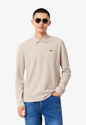 Polo beige a maniche lunghe con colletto, caratterizzata da un piccolo logo Lacoste verde sul petto a sinistra, abbinata a un paio di jeans blu.