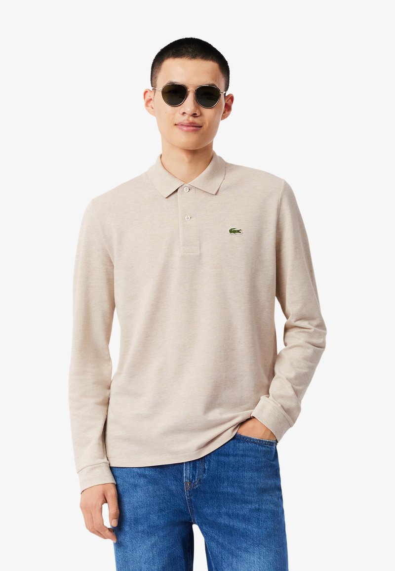 Polo beige a maniche lunghe con colletto, caratterizzata da un piccolo logo Lacoste verde sul petto a sinistra, abbinata a un paio di jeans blu.