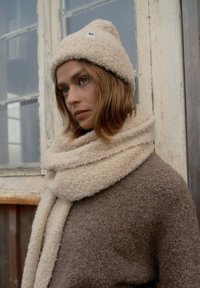 Cappello in lana beige con un'etichetta decorativa, abbinato a una morbida sciarpa coordinata e a un cappotto marrone texturizzato. Ritratto in primo piano su uno sfondo rustico.