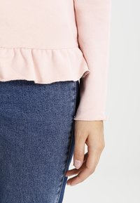 Top lungo rosa a maniche, con orlo arricciato, realizzato in morbido tessuto. Abbinato a jeans blu, che mostrano bordi delle maniche sfrangiati e dettagli fatti a mano.