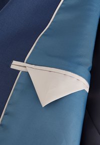 Doublure de blazer bleu et crème avec une boucle en tissu bleu maintenant le rabat de la poche intérieure crème contre l’intérieur bleu.