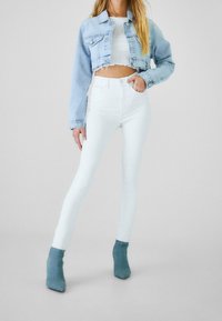 Veste courte en denim bleu clair portée sur un crop top blanc, associée à un jean skinny blanc taille haute et des bottines bleu sarcelle.
