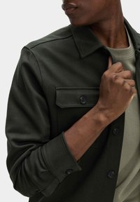 Giacca camicia in verde scuro a spina di pesce con colletto, tasca sul petto e chiusura con bottoni sui polsini. Indossata sopra una camicia verde chiaro.